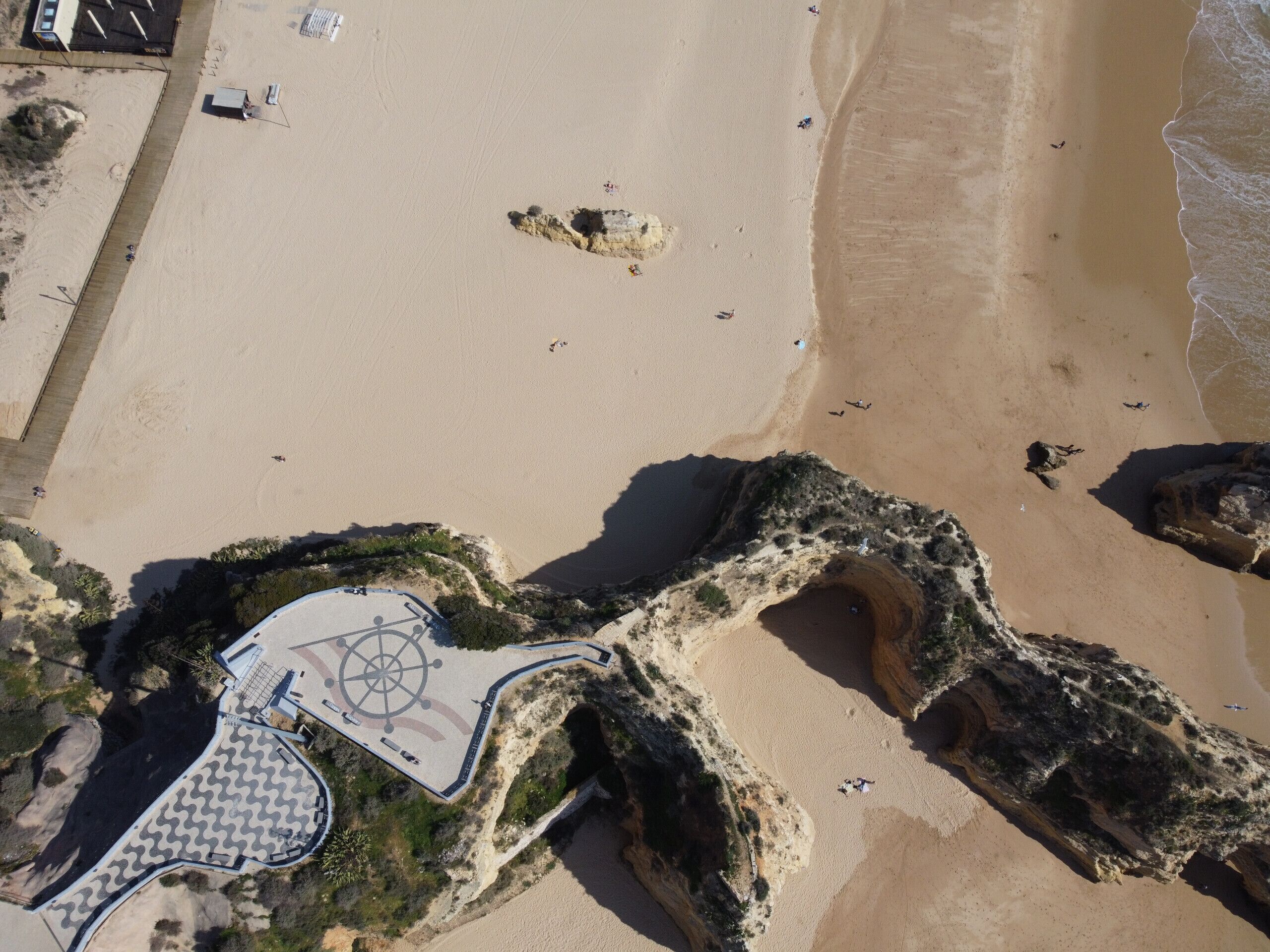 An aerial view of a beach with a paved area featuring a circular design, rocky formations, and a sandy shoreline.