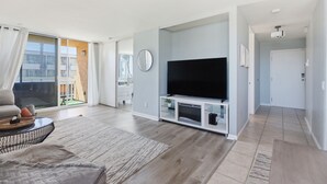 A modern living room with a flat-screen TV, a sofa, a coffee table, and a large window with curtains. - Ocean View Oasis in Pacific Beach 2BR 2BA Balcony Pool  Monthly Stay (San Diego)