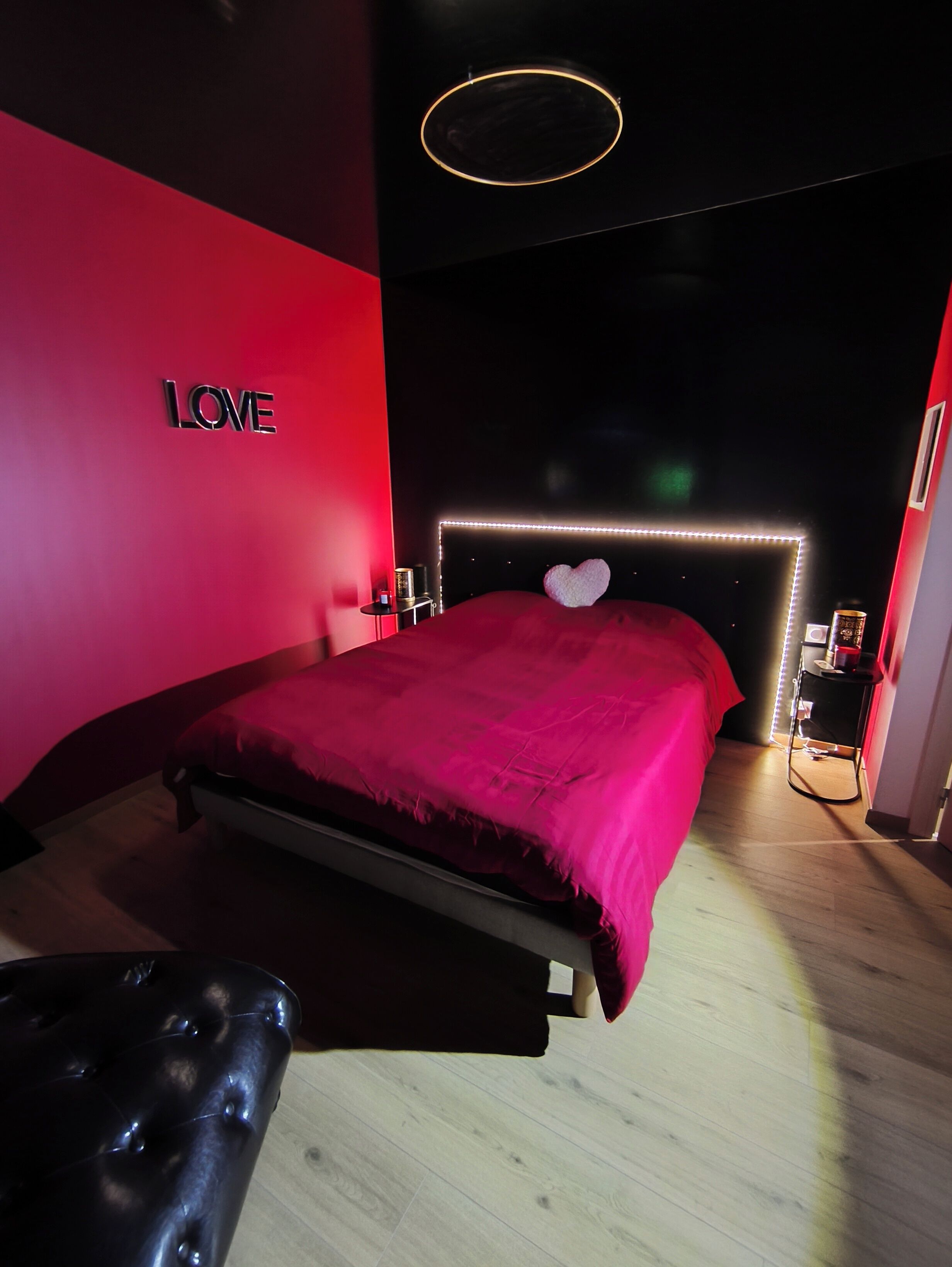 Un dormitorio moderno con una pared de acento roja, una cama con un cojín con forma de corazón y una mesita de noche con una lámpara.