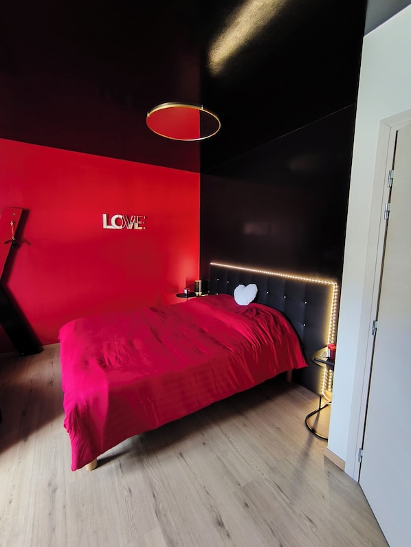 Une chambre moderne avec un mur d’accent rouge, un lit agrémenté d’un coussin en forme de cœur et un luminaire fixé au mur.