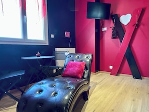Une pièce avec un fauteuil en cuir noir, une petite table sur laquelle se trouve une pomme, un téléviseur accroché au mur et un objet décoratif en forme de cœur.