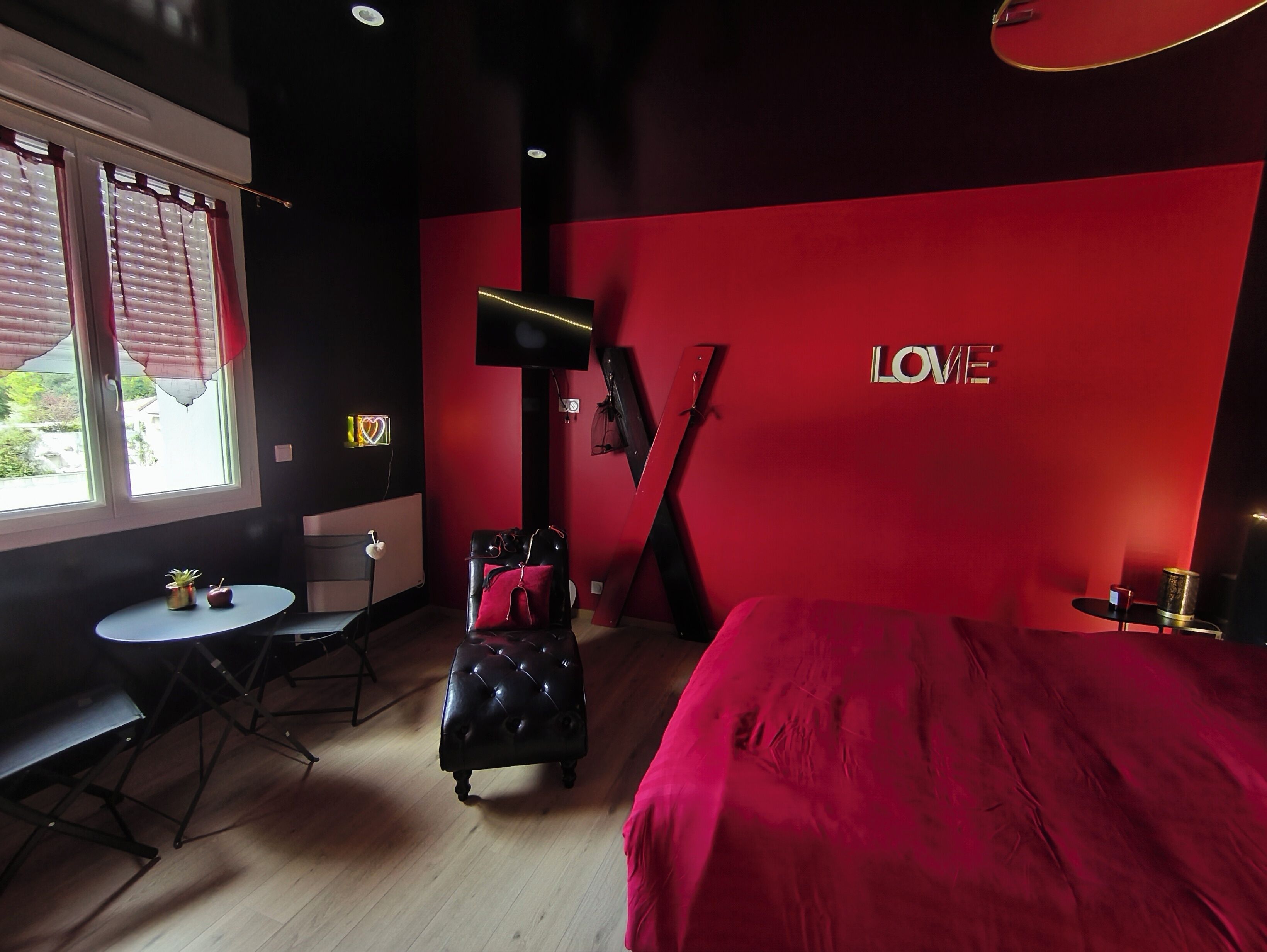 A modern hotel room with a red accent wall, a bed, a desk, a chair, a small table, and a TV mounted on the wall.
