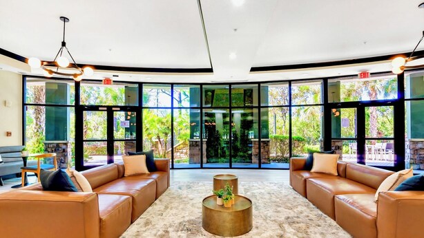 A spacious lobby with leather sofas, a central coffee table, and large windows offering a view of greenery outside. - Upstay Pet-friendly Condo With Pool (Winter Garden)