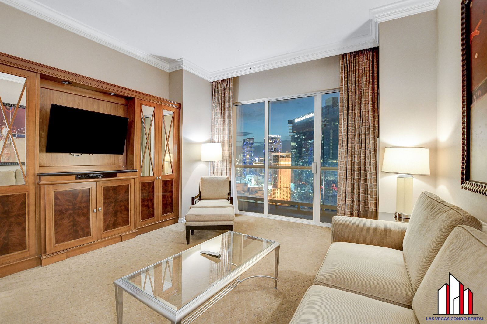 A modern living room with a flat-screen TV, wooden cabinets, a glass coffee table, a beige sofa, and a large window with a city view.
