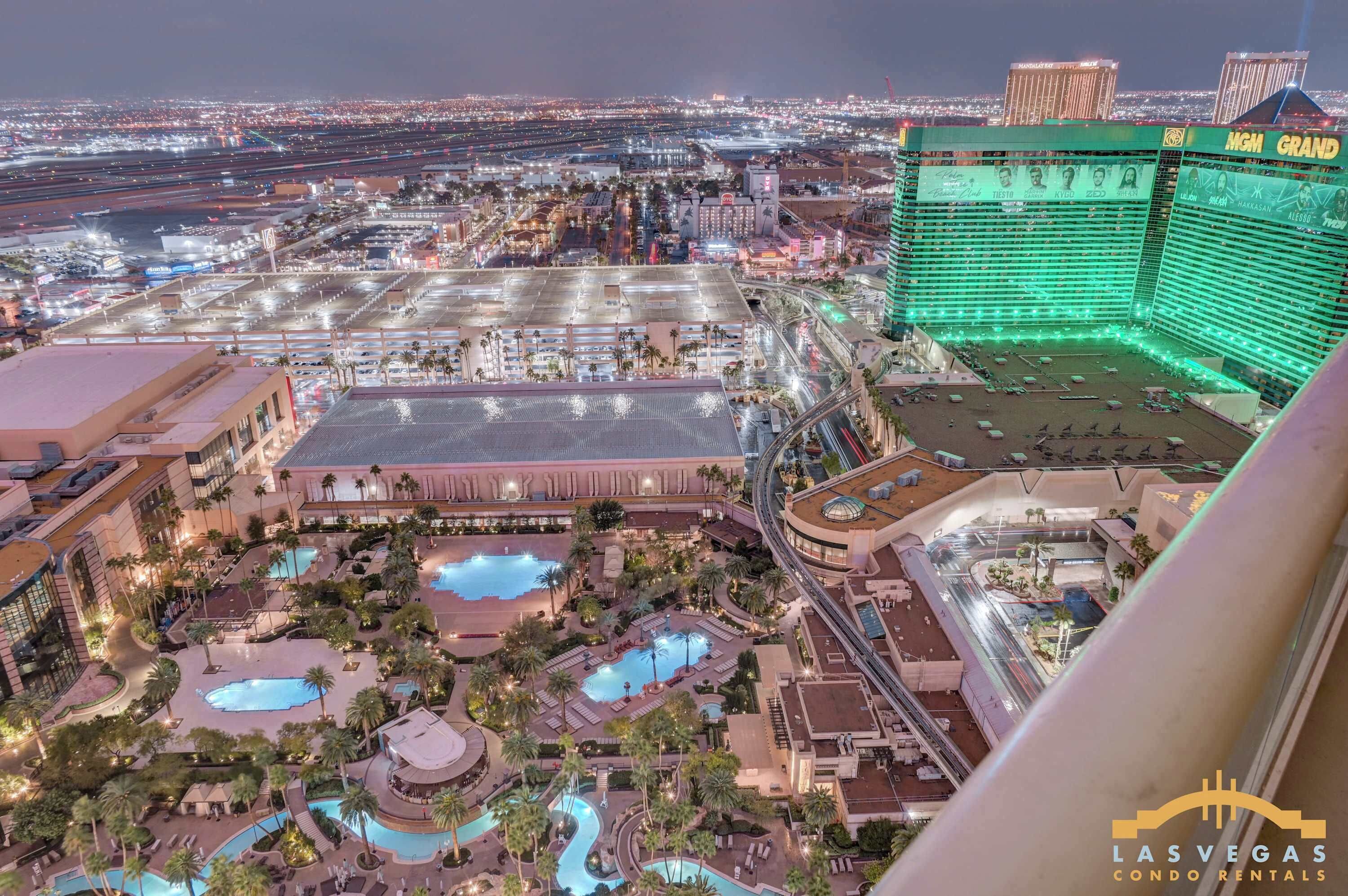 A hotel with multiple swimming pools, a large parking area, and a view of the surrounding cityscape.
