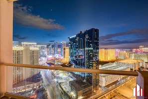 A nighttime cityscape with high-rise buildings, a view of the streets below, and a balcony railing.
