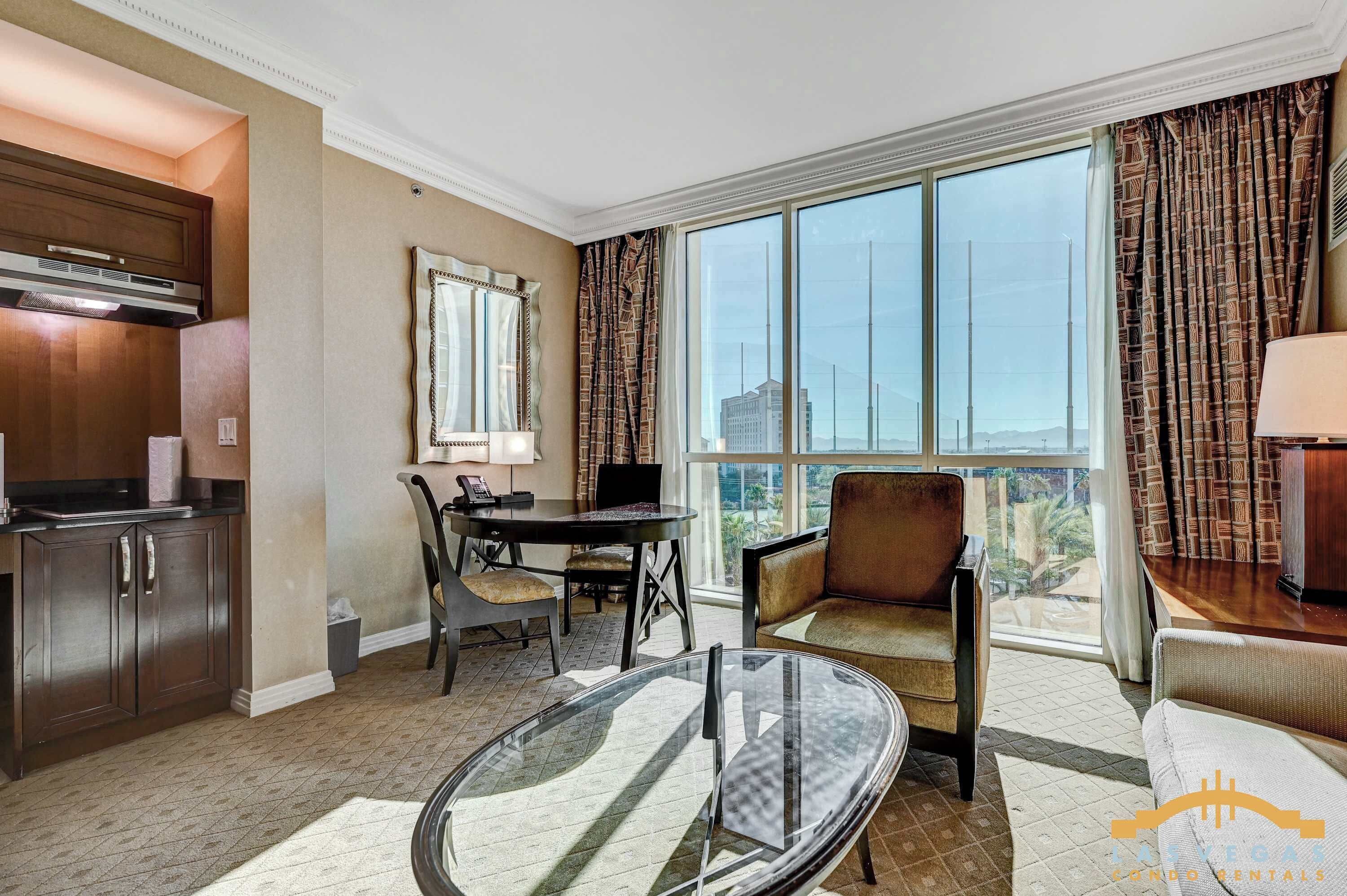 A hotel room with a dining table, chairs, a sofa, a glass table, a kitchen area, and large windows with curtains.