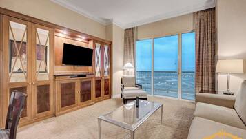A hotel living room with a large TV, a glass coffee table, a sofa, and a view of the ocean.