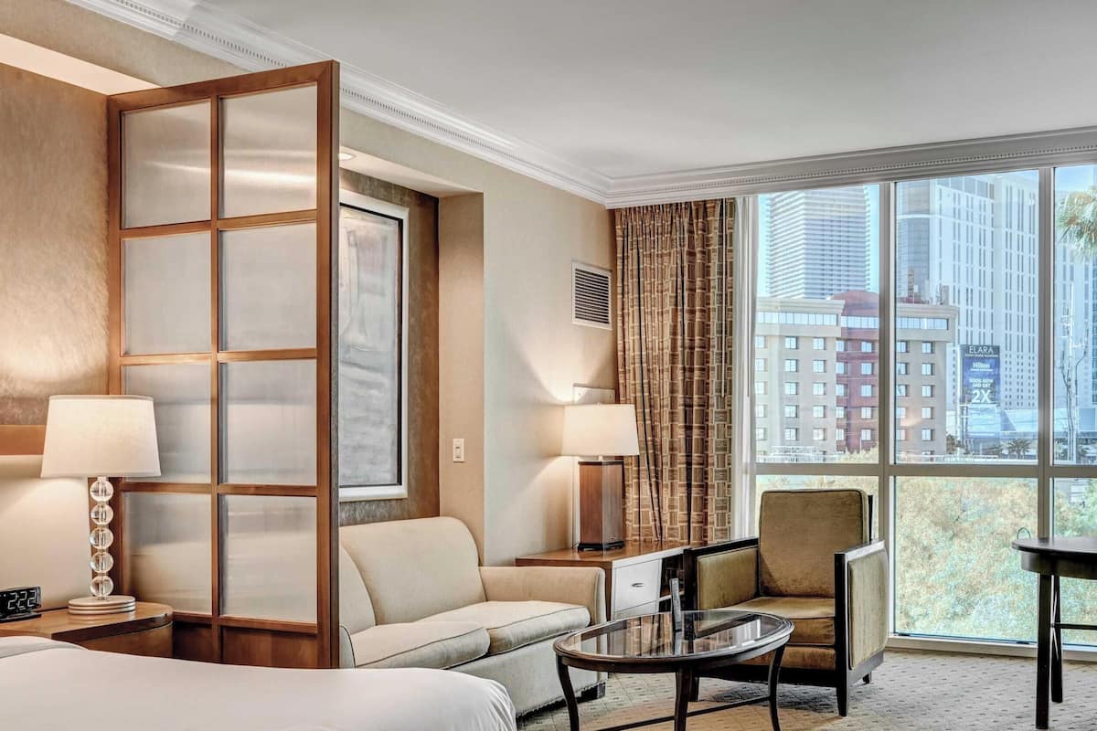 Habitación de hotel con cama, sofá, una mesita, dos sillas, un ventanal con vista a edificios y un separador decorativo.
