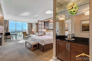 Habitación de hotel con cama, escritorio, silla, balcón con vistas y zona de cocina.