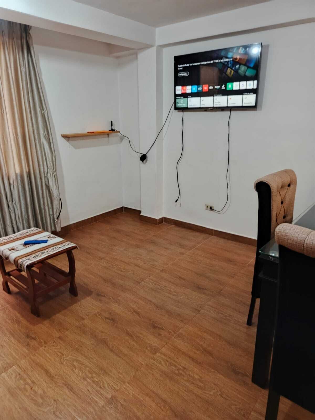 A room with a wooden floor, a television mounted on the wall, a small wooden table, and a chair.