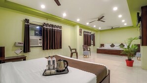 Een hotelkamer met een bed, nachtkastje, gordijnen, een raam met airconditioning, een plafondventilator, een potplant en een bureau met stoel.