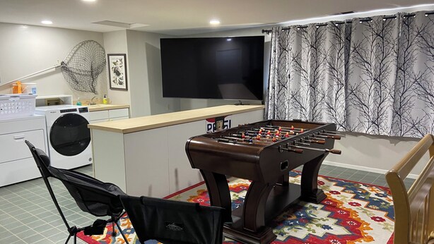 Una habitación con mesa de futbolín, lavadora, televisor y ventana con cortinas.