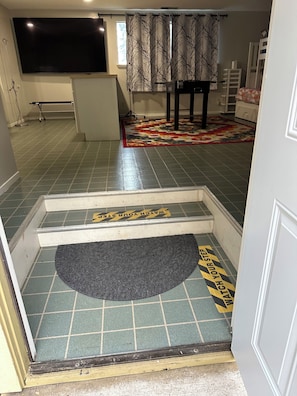 A hotel room with a tiled floor, a flat-screen TV mounted on the wall, a bed, a bedside table, and a window with curtains.