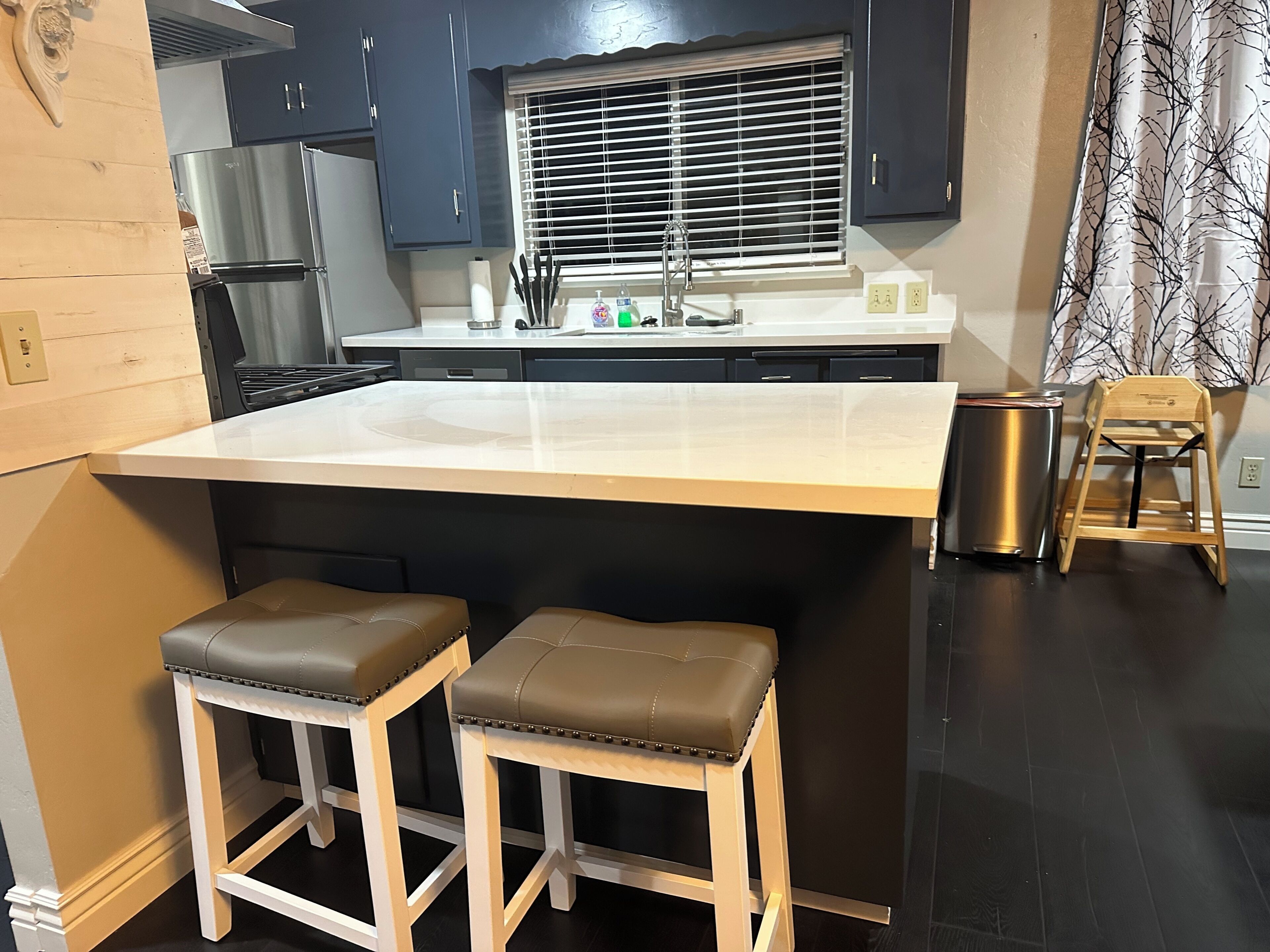 A kitchen with a large island, two stools, a sink, and a window with blinds.