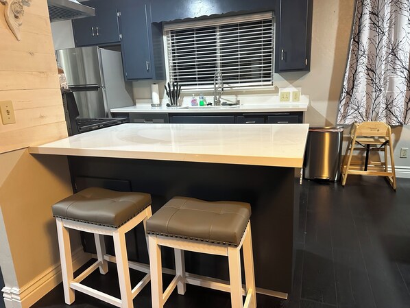 Una cocina con una isla grande, dos taburetes, un fregadero y un ventanal con persianas.