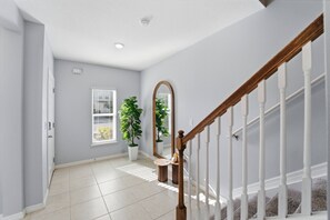A hallway with a wooden staircase, a white door, a potted plant, a mirror, and a window with blinds. - 3BR Family Home at Fiesta Key Near Parks (Kissimmee)