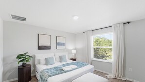 Ett modernt sovrum med en stor säng, ett sängbord, en liten avlastningsyta, ett fönster med gardiner och en krukväxt.
