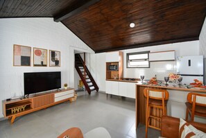 A modern kitchen with a wooden ceiling, white brick walls, a built-in TV, and a dining area with wooden chairs. - Casa na Praia dos Ingleses -NIN019 (Florianópolis)
