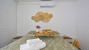 Ein Zimmer mit einem Bett, einer Wandklimaanlage, einer gewebten Wanddekoration, einem kleinen Holztisch mit einer Vase und Obst, und ordentlich gefalteten Handtüchern.