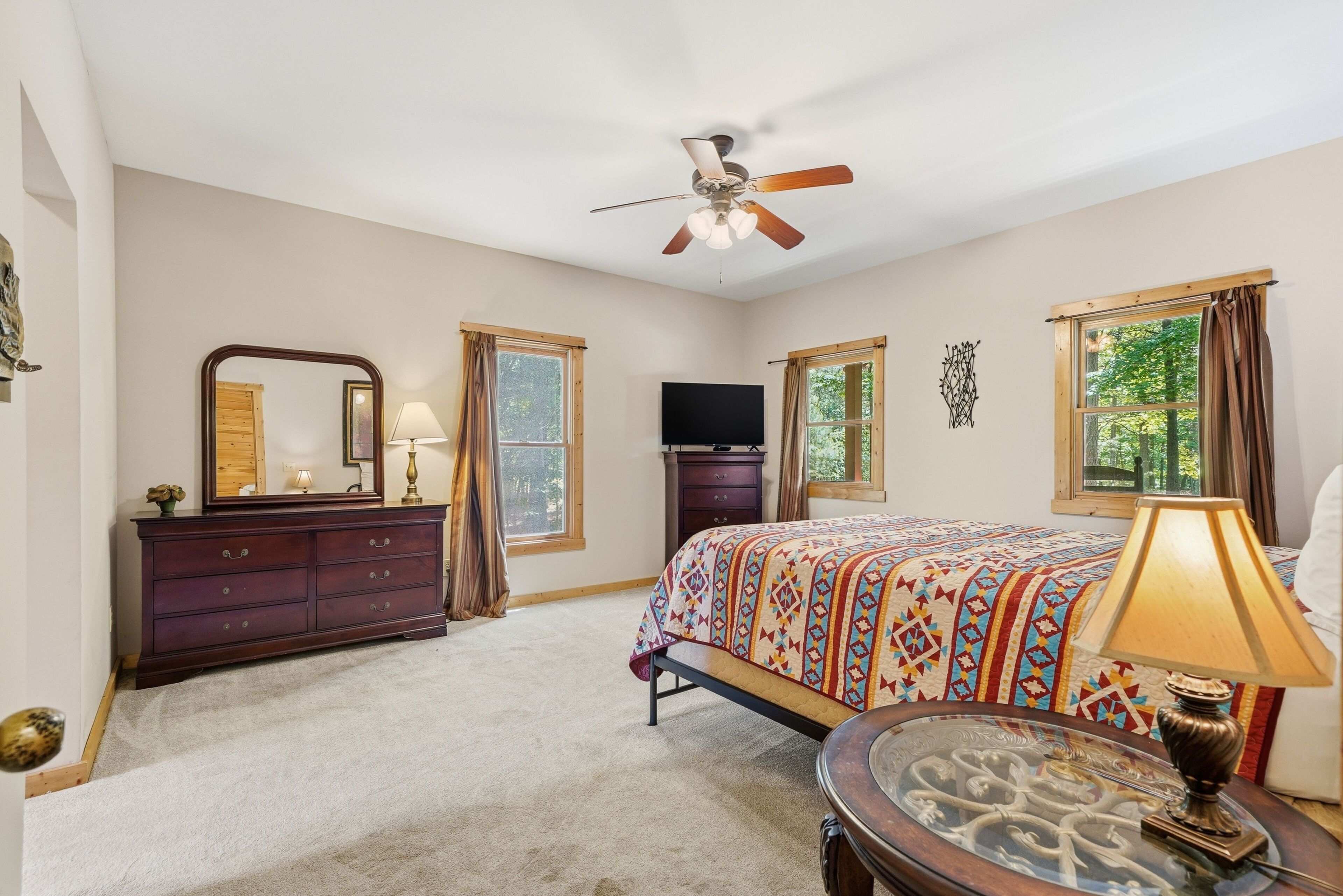 A bedroom with a bed, dresser, television, ceiling fan, and windows.