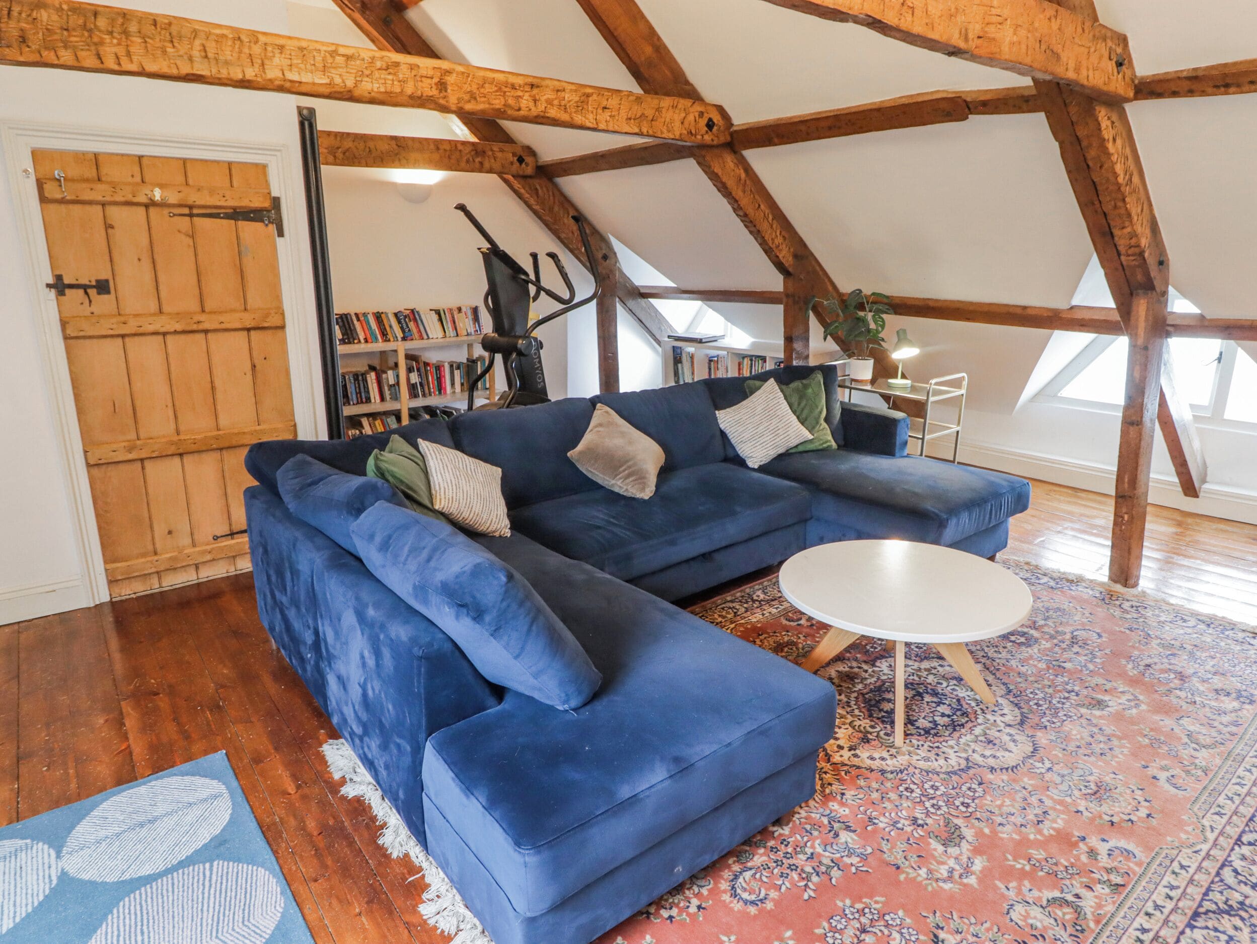 Una habitación estilo loft con vigas de madera a la vista, un sofá seccional azul, una mesita blanca y un tapete estampado.