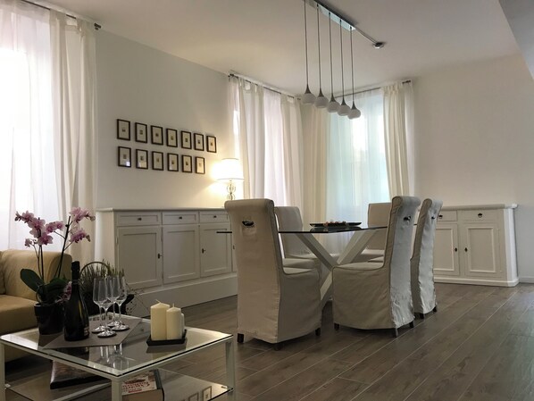 A modern living room with a glass coffee table, beige armchairs, a dining table with chairs, white curtains, and framed pictures on the wall. - In the historic center of Bellagio, a large, cozy, and bright apartment (Bellagio)