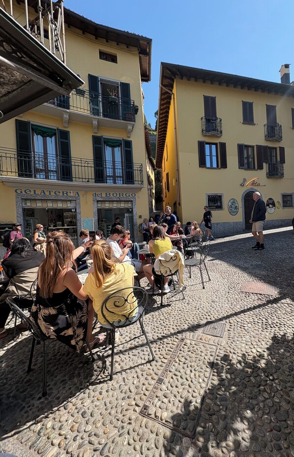 Un café al aire libre con mesas y sillas, una calle adoquinada y edificios con balcones.