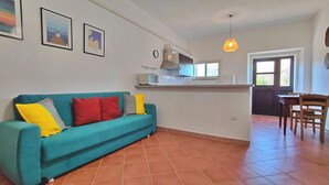 Una sala de estar con un sofá color turquesa, un comedor con una mesa y sillas de madera, una cocina con estufa y ventana, y un reloj en la pared.