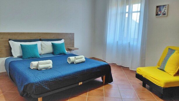 Una cama con ropa de cama azul, almohadas blancas y toallas enrolladas. Hay una silla amarilla con un cojín a rayas.