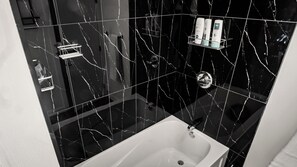 A bathroom with a white bathtub, black marble tiles, and a shelf with toiletries. - Luxury Comfort Stay in Keswick | 2Bedrooms- 1Bath (Edmonton)