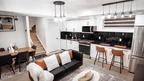 A modern kitchen with a stove, microwave, and refrigerator. There is a dining area with a table and chairs, and a sofa with cushions. A staircase is visible in the background. - Luxury Comfort Stay in Keswick | 2Bedrooms- 1Bath (Edmonton)
