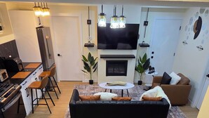 En moderne stue med sofa, spiseplads med stole, køkken med komfur og et fjernsyn.