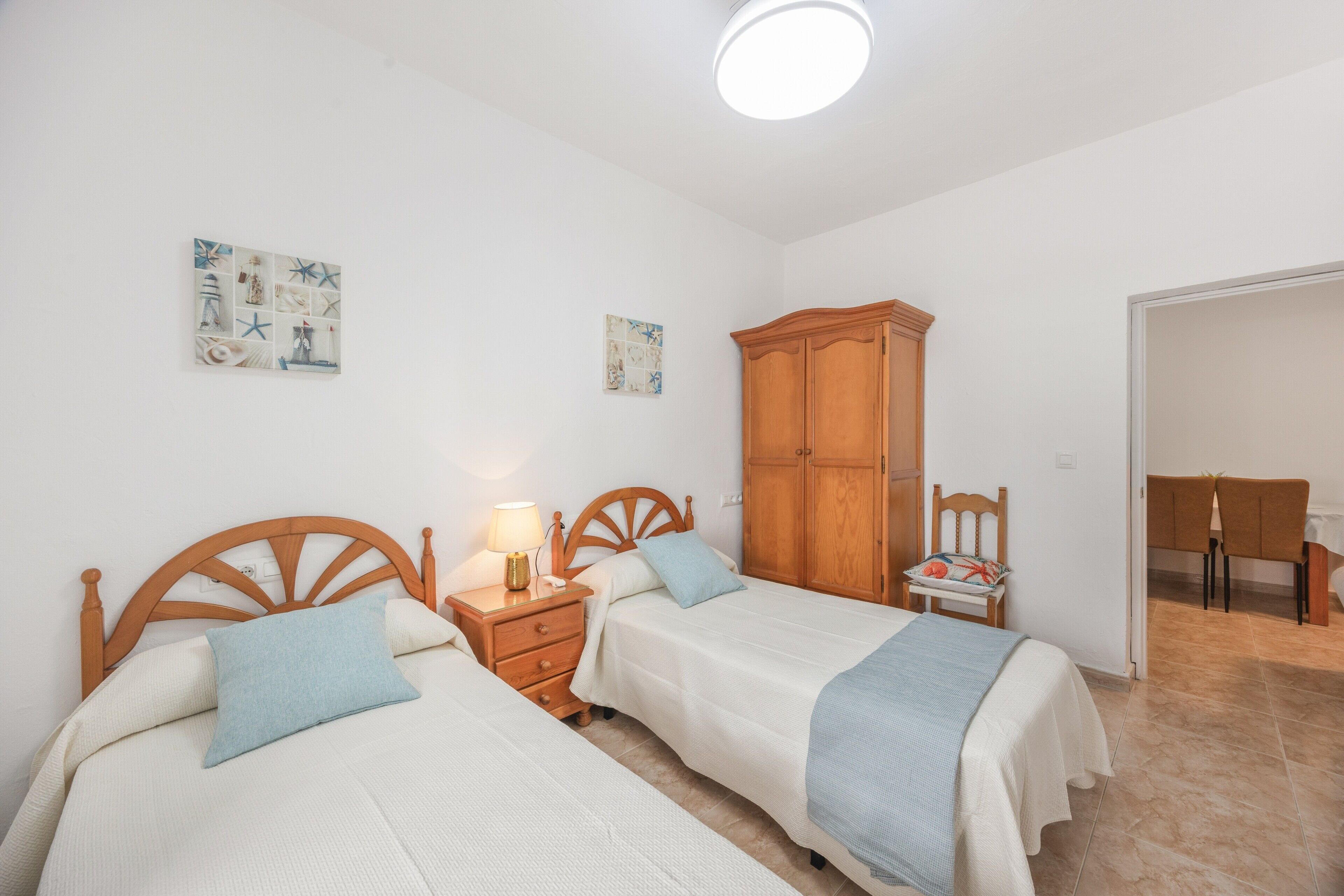 Une chambre avec deux lits, une armoire en bois, une table de chevet avec une lampe, et un coin repas avec des chaises.