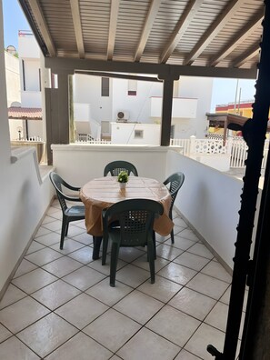 Een overdekt balkon met een ronde tafel en vier stoelen, een bloempot en een uitzicht op gebouwen op de achtergrond.