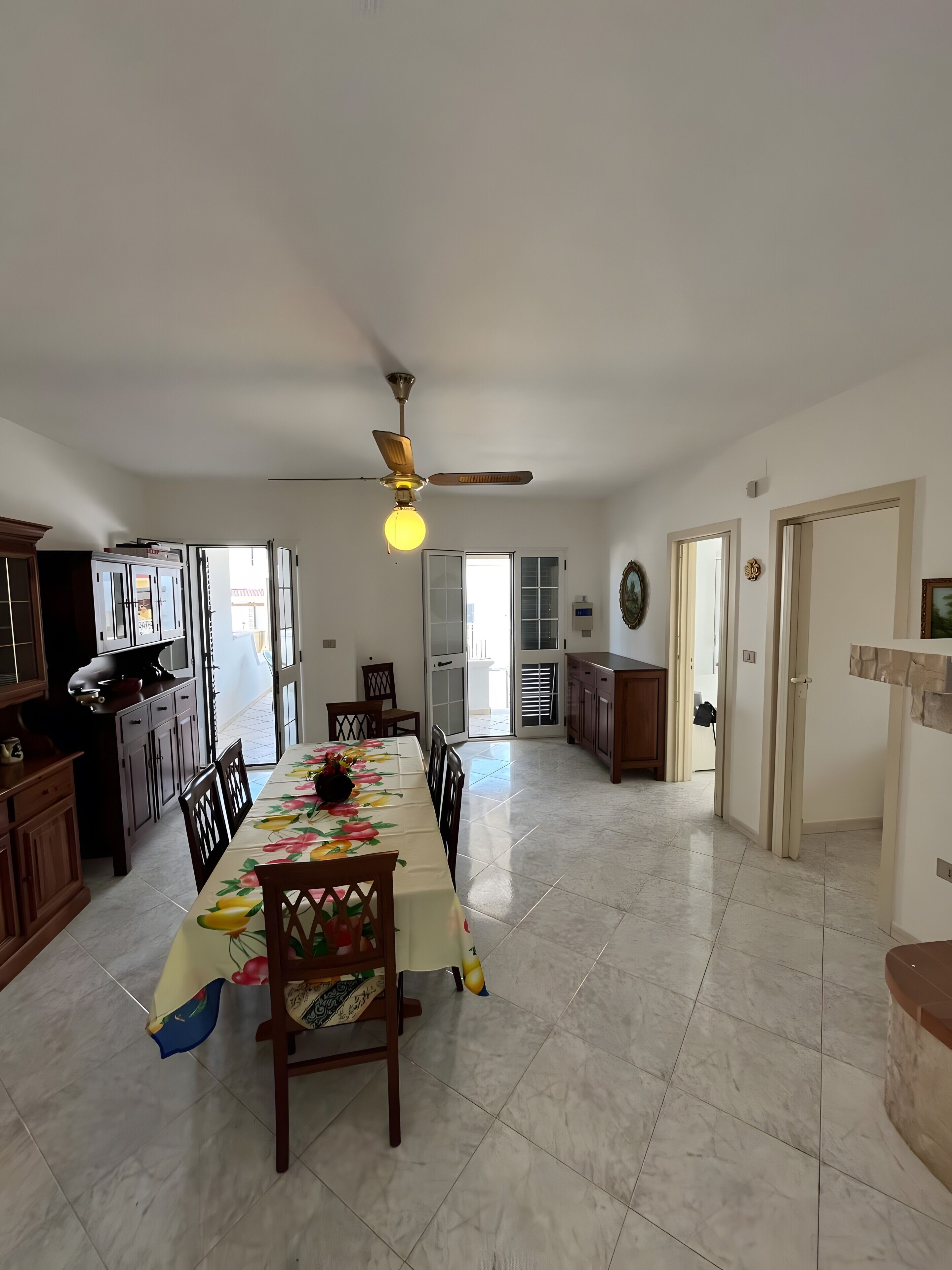 Uma sala de jantar ampla, com mesa posta para quatro pessoas, ventoinha no teto e cozinha equipada com armários e micro-ondas.