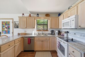 A kitchen with wooden cabinets, a microwave, a stove, a sink, and a window with a view of trees. - Cozy Murphy Cabin w/ Mountain Views (Murphy)