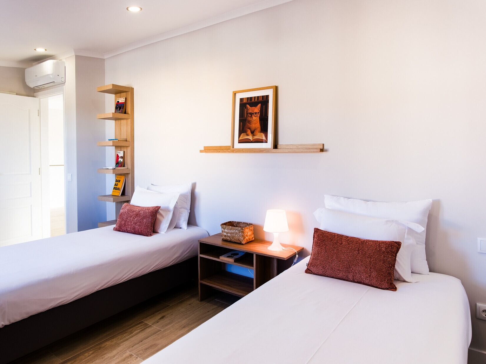 A room with two beds, a shelf with books, a framed picture, a lamp, and a basket.