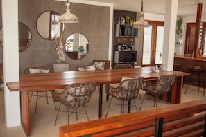 A modern kitchen with a long wooden island, wicker chairs, and pendant lighting.