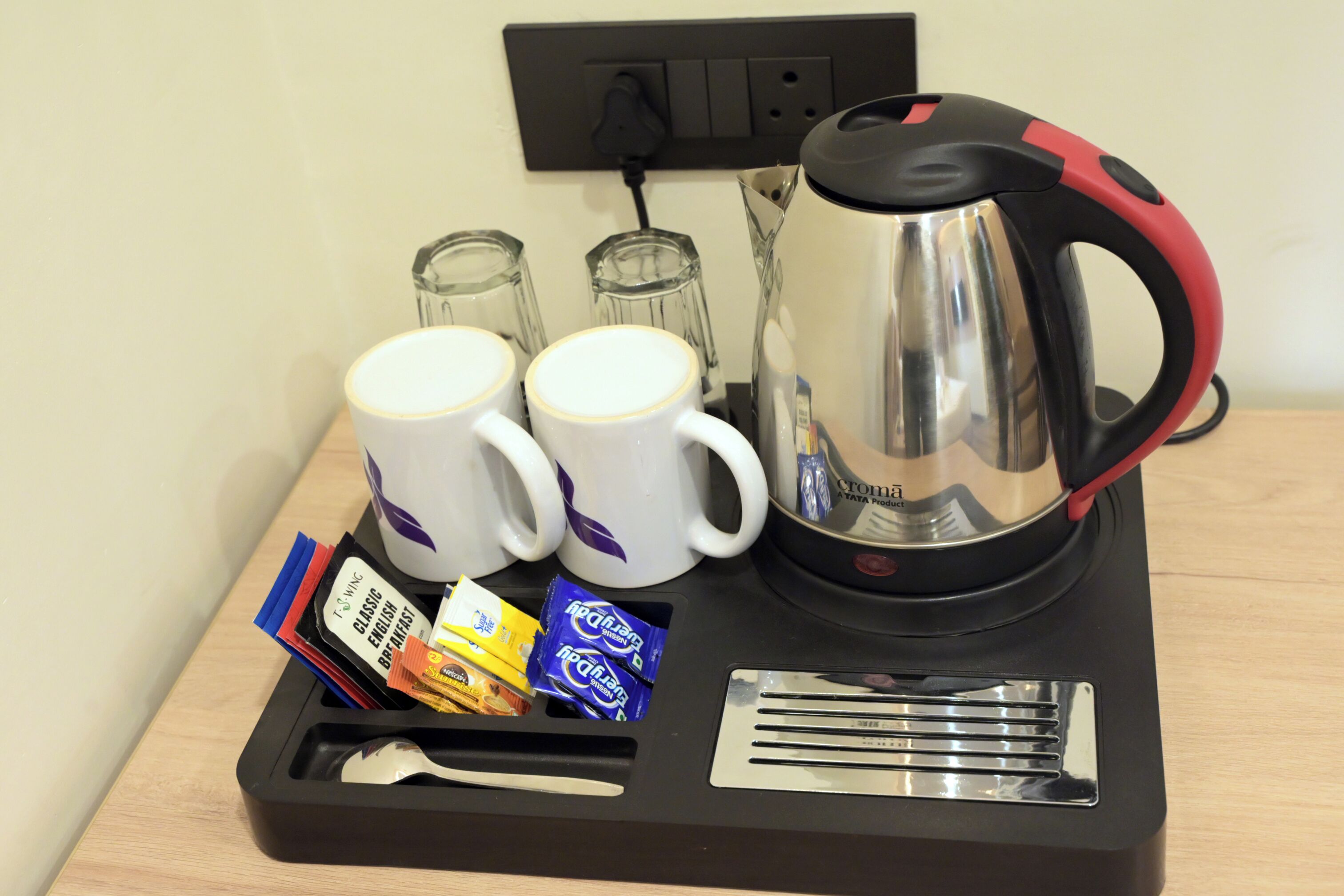 Een dienblad met twee witte mokken, een zilveren elektrische waterkoker, een lepel en wat snacks.