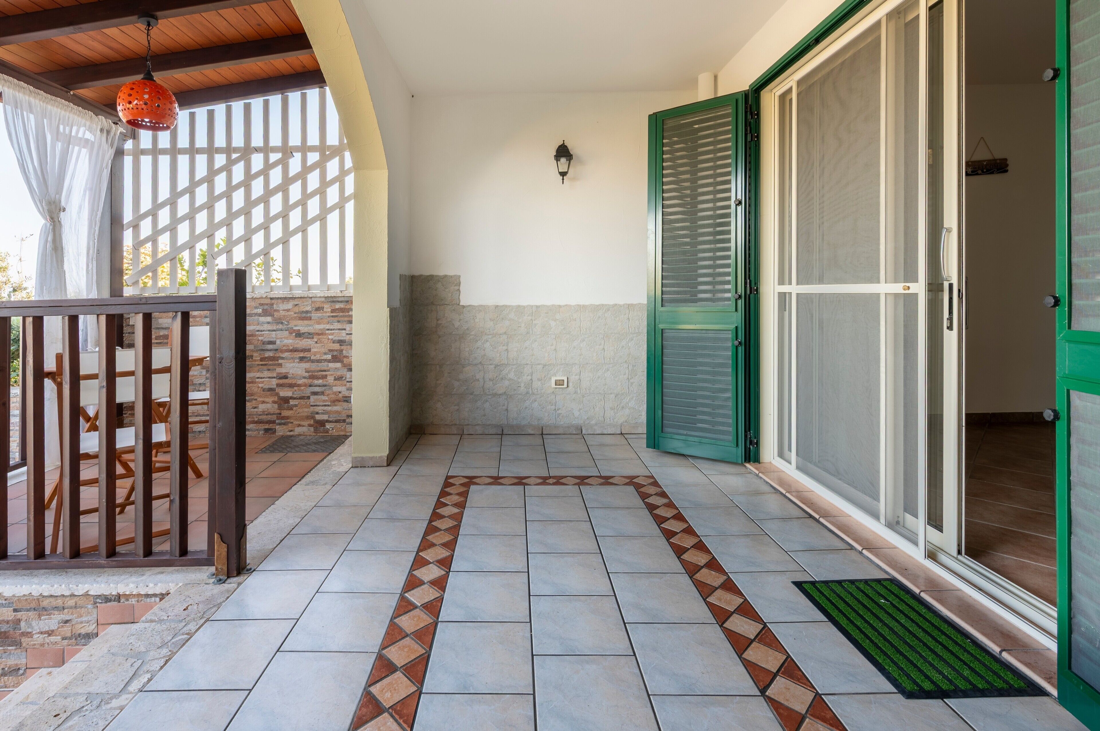 Un'area patio coperta con pavimento piastrellato, recinzione in legno e una porta con persiane verdi.