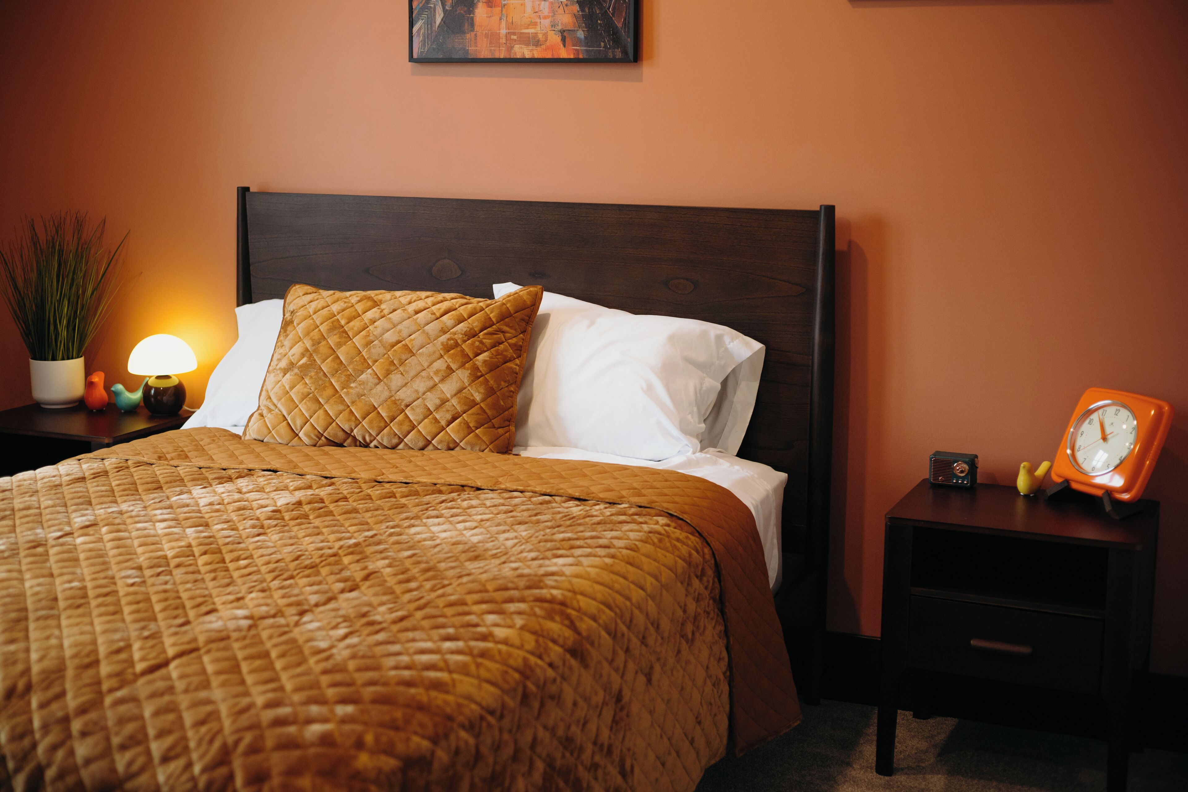 Una cama bien hecha con cubrecama acolchada, una mesita de noche con un reloj y un cuadro colgado en la pared.