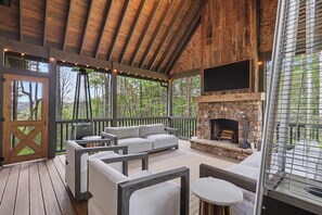 A covered outdoor seating area with a stone fireplace, wooden deck, and a view of the surrounding forest.