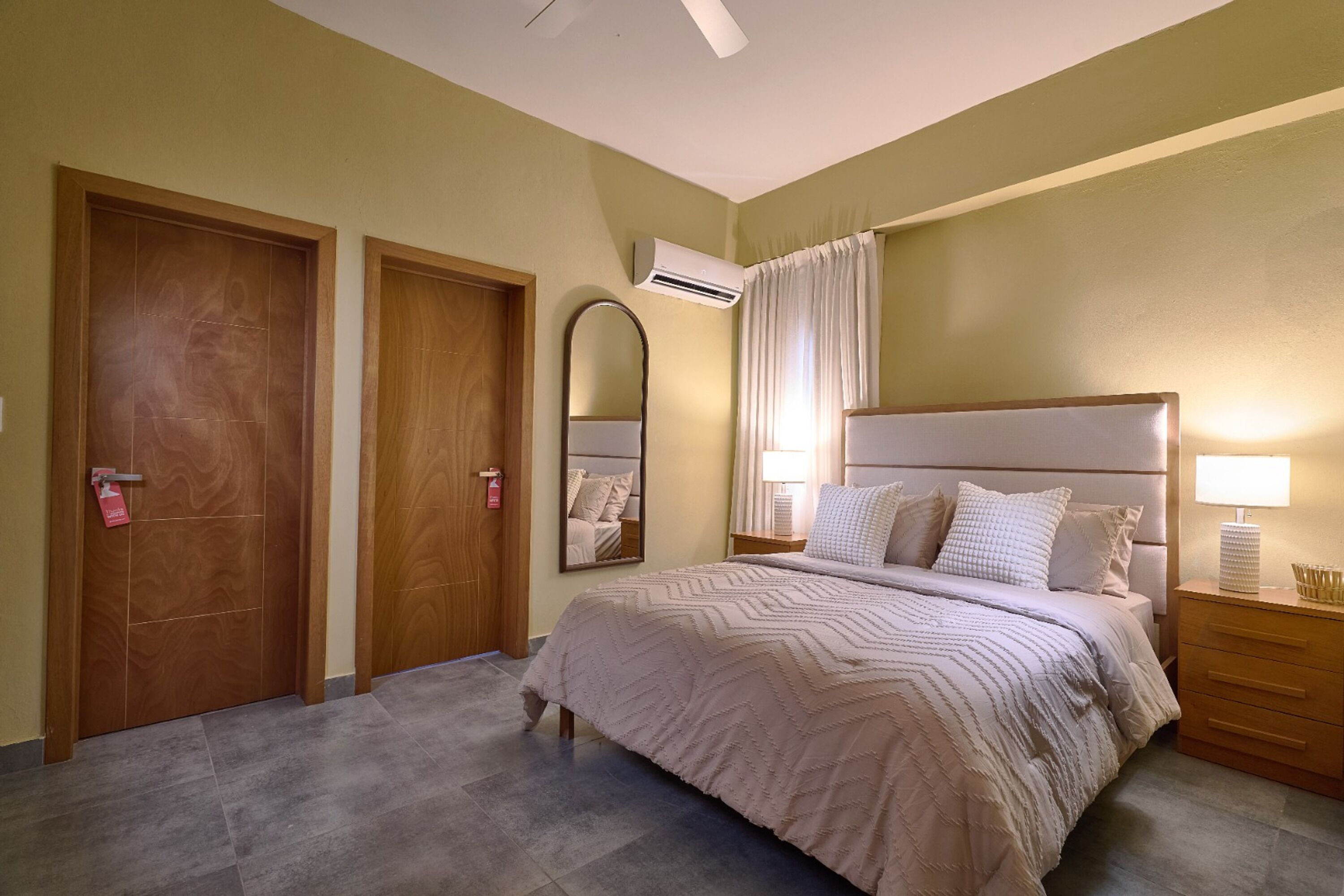 Une chambre d’hôtel comprenant un lit, deux portes, un appareil de climatisation, un miroir, une table de chevet et une lampe.