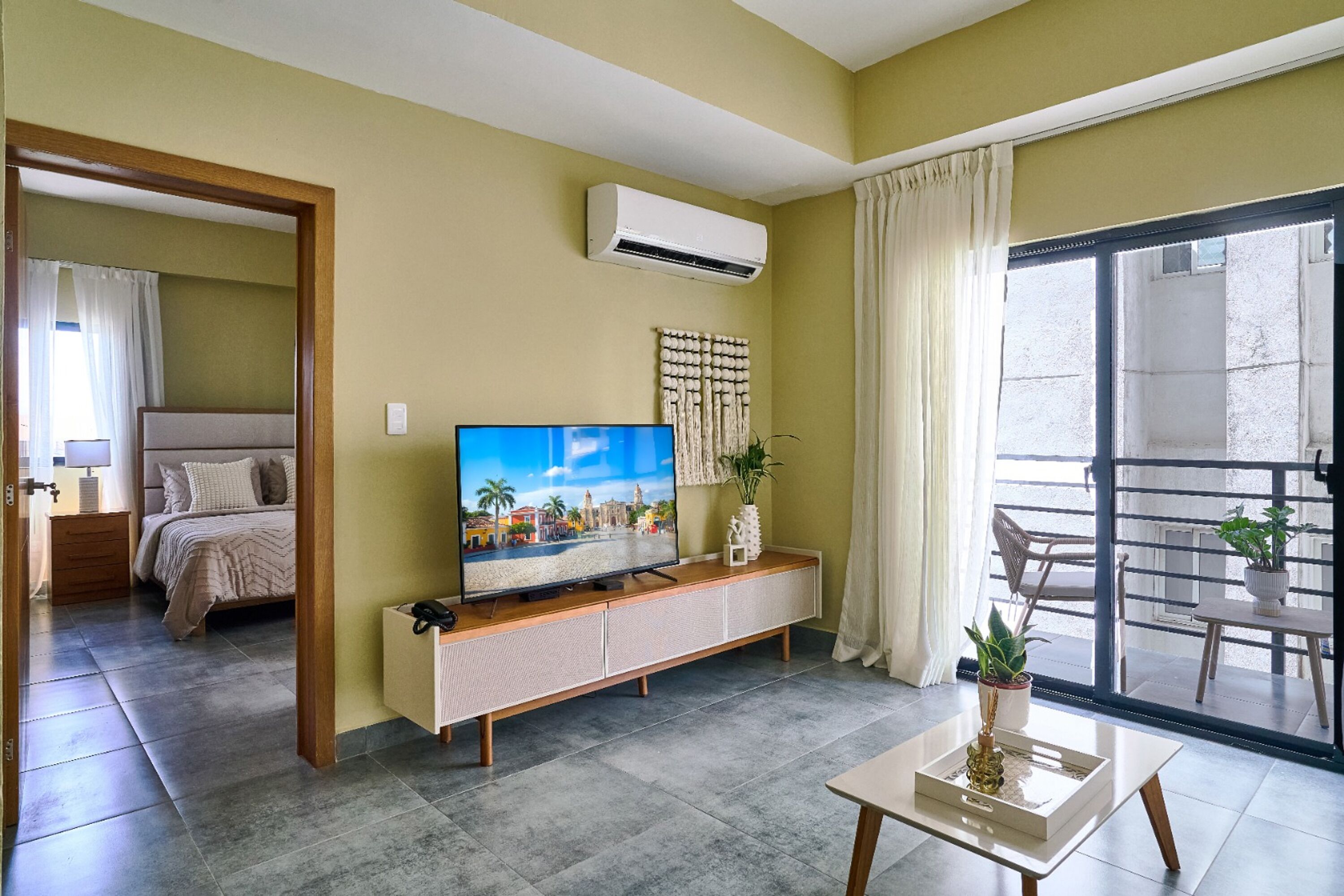 Een moderne woonkamer met een flatscreen televisie, een houten kast, een kleine tafel met een vaas, en een schuifdeur die leidt naar een balkon met bloempotten.