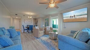 A spacious living room with a large window, a ceiling fan, and a dining area with blue chairs. - Sunkissed Seaside by Avantstay Walk to Beach + Boardwalk Pool + Tennis + Basketball Access (Port Aransas)