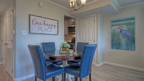 A dining area with a round table, four blue chairs, a chandelier, a kitchen in the background, and a painting of a bird on the wall. - Sunkissed Seaside by Avantstay Walk to Beach + Boardwalk Pool + Tennis + Basketball Access (Port Aransas)
