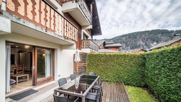 Ein moderner Essbereich im Freien mit einem Glastisch und Stühlen, einer Holzterrasse und Blick auf eine Berglandschaft.