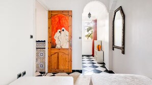 A hotel room with a checkered floor, a wooden door with a painting, a white bed, a mirror, and a patterned cabinet. - Lovely 2-El Jadida City Center Apt – Ideal Spot, Beach View & WiFi (El Jadida)