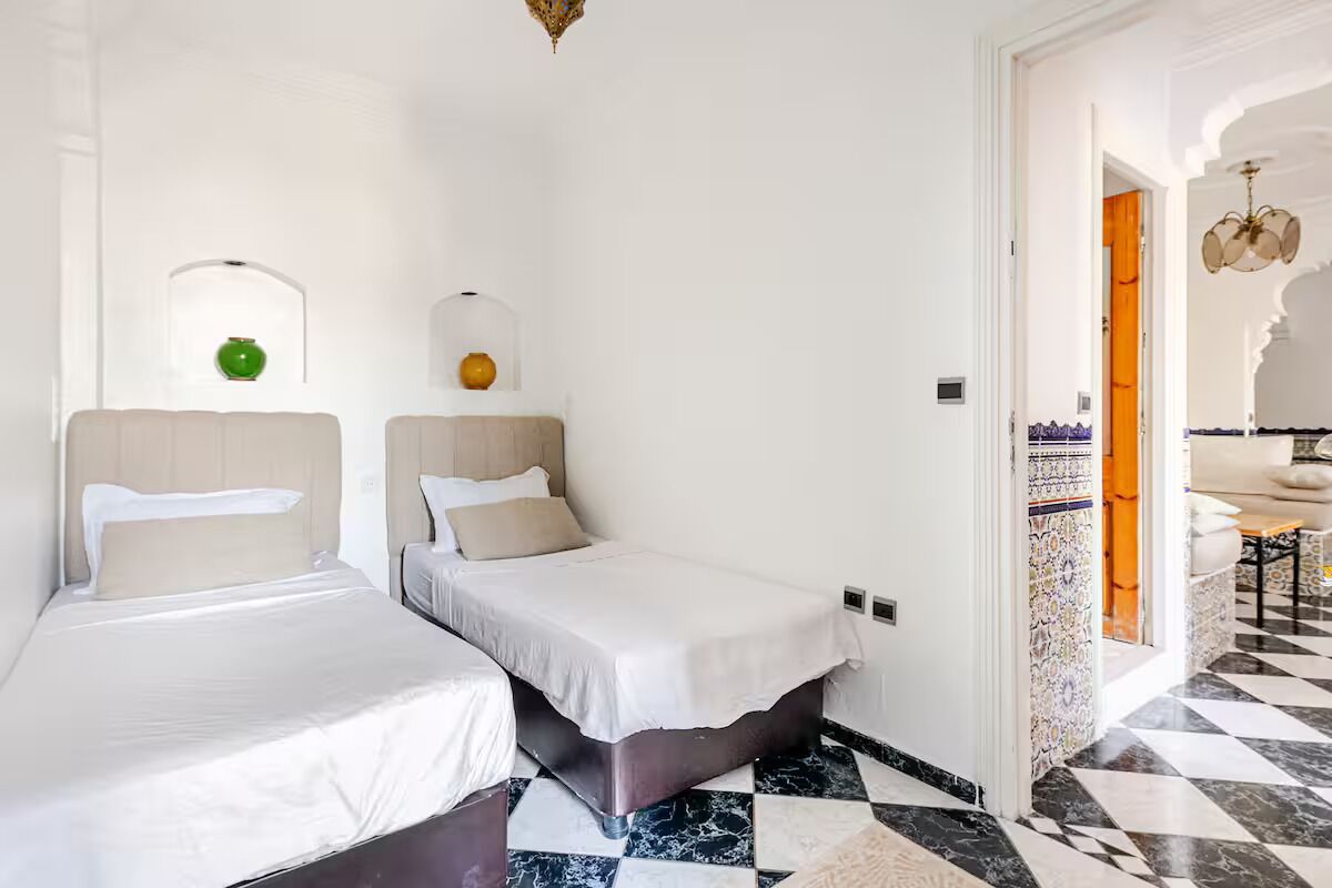 Una habitación de hotel con dos camas, un suelo a cuadros, una mesita y un baño visible a través de una puerta abierta.